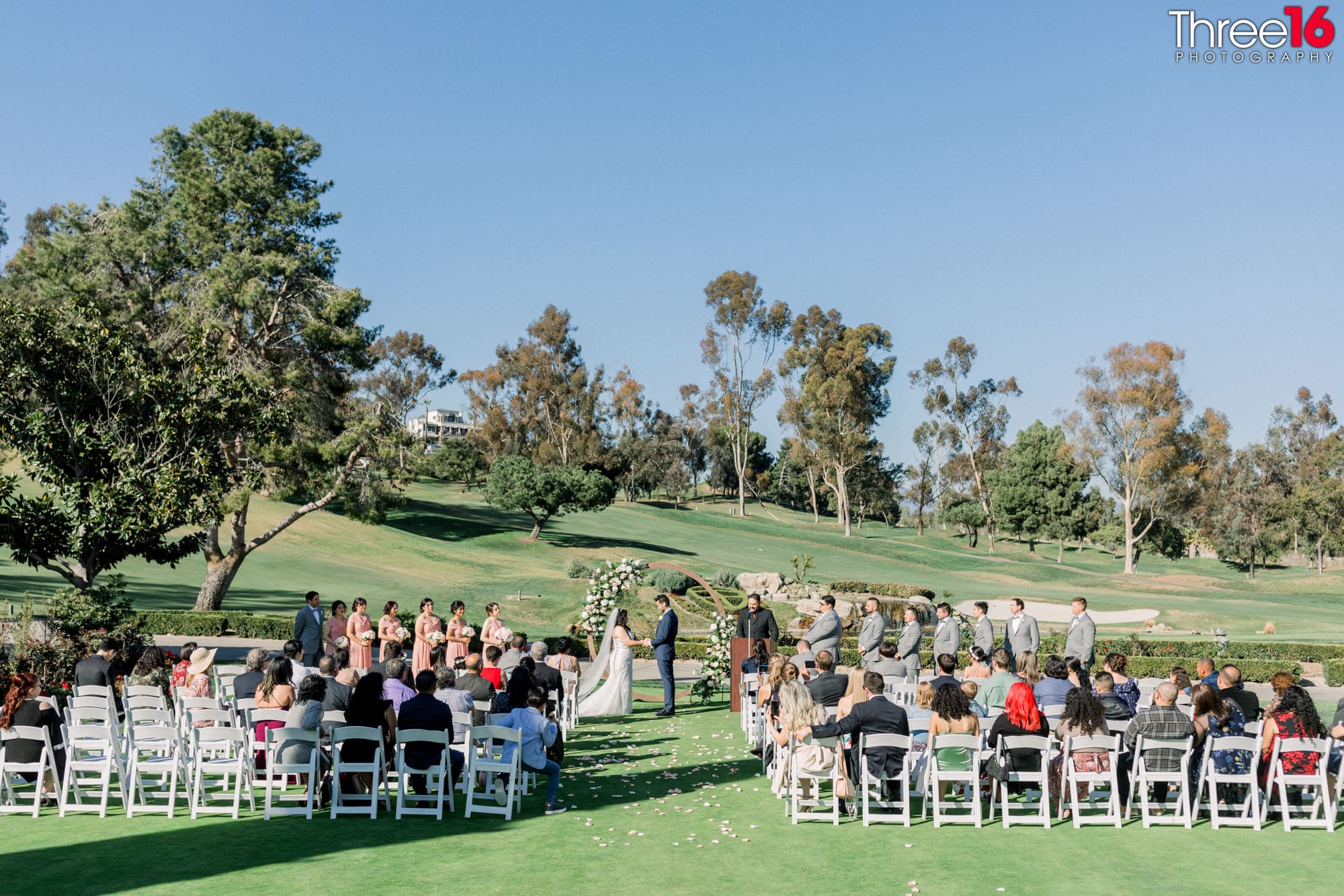 Bride and Groom face each other and hold hands while taking their vows on the green grass of the Marbella Country Club wedding venue in San Juan Capistrano.