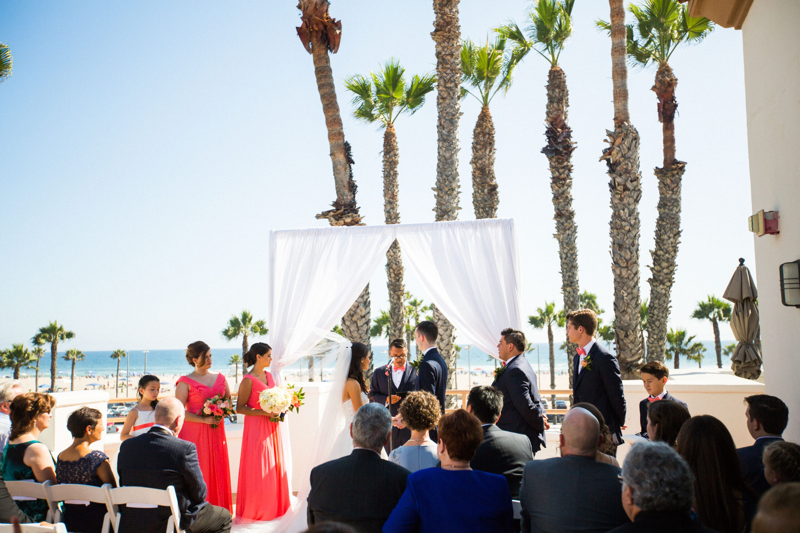 Bride and Groom take their vows during their Hilton Waterfront Beach Resort wedding venue in Huntington Beach as wedding party and guests look on.