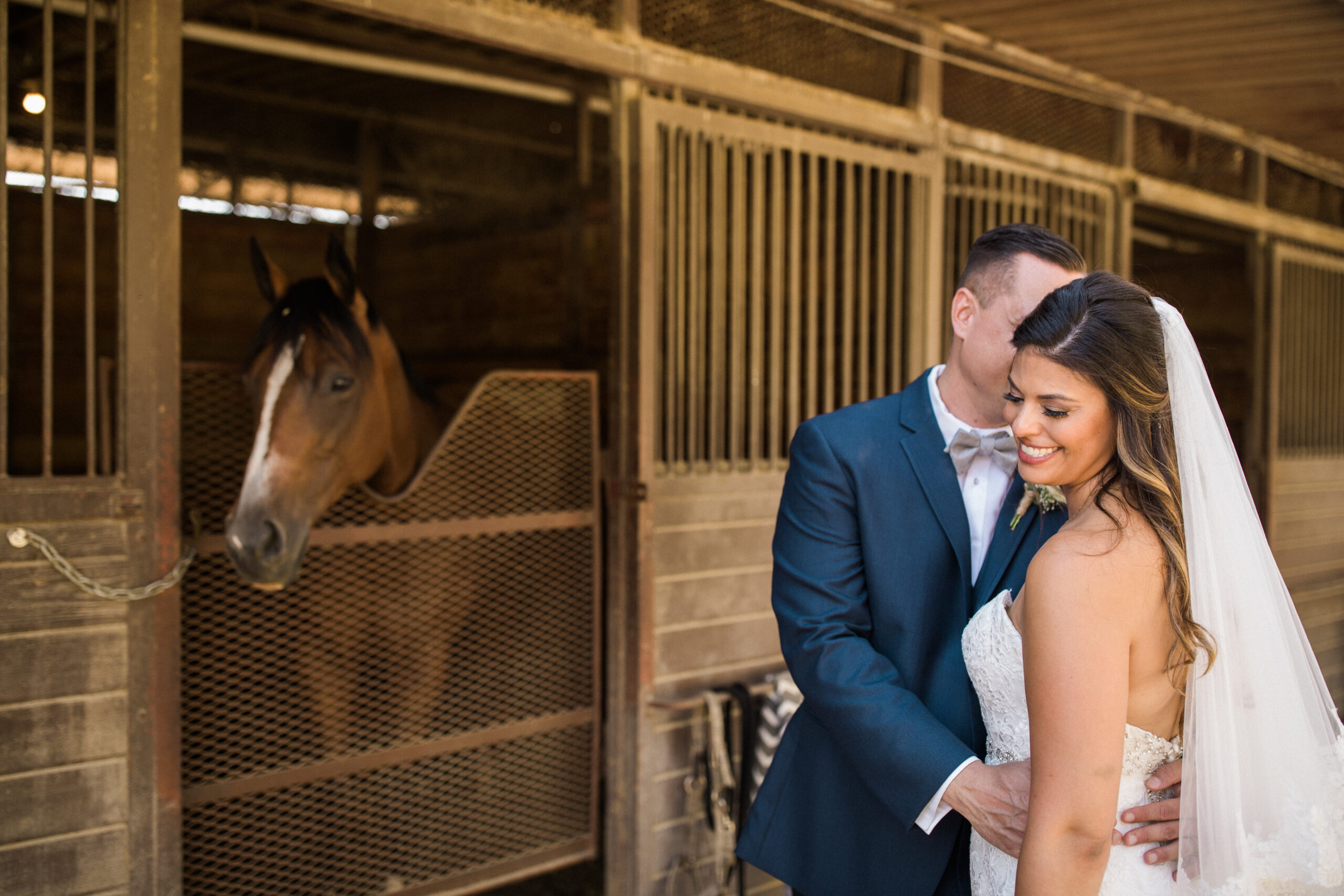 Groom whispers into his smiling Bride's ear in front of a horse in a stable at The Red Horse Barn wedding venue in Huntington Beach.